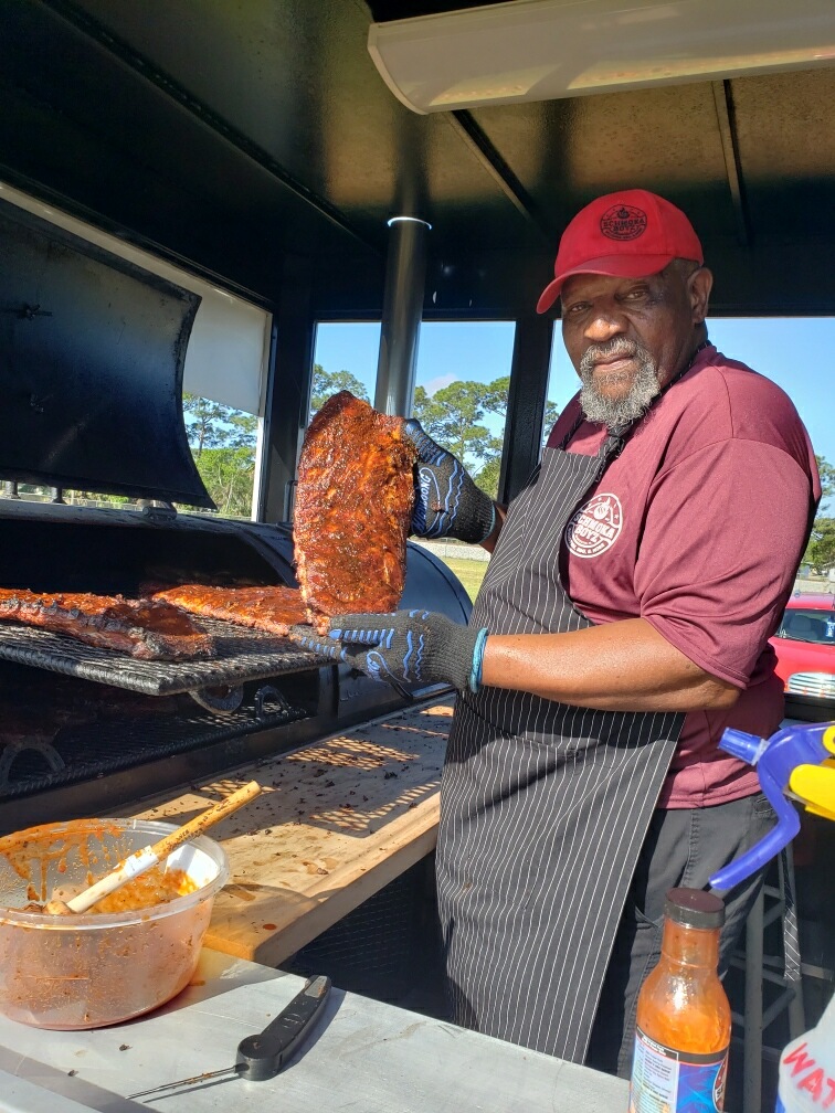 Man holding rack of ribs at Schmoka Boyz BBQ, with more ribs on the grill.